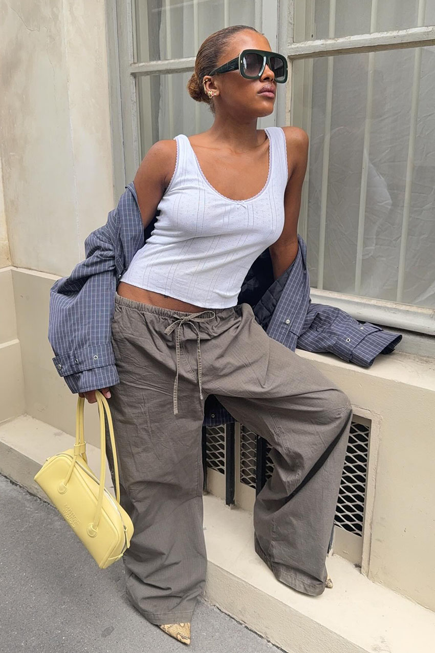 A woman wearing a white tank top, plaid button down, grey drawstring pants, and a yellow leather handbag