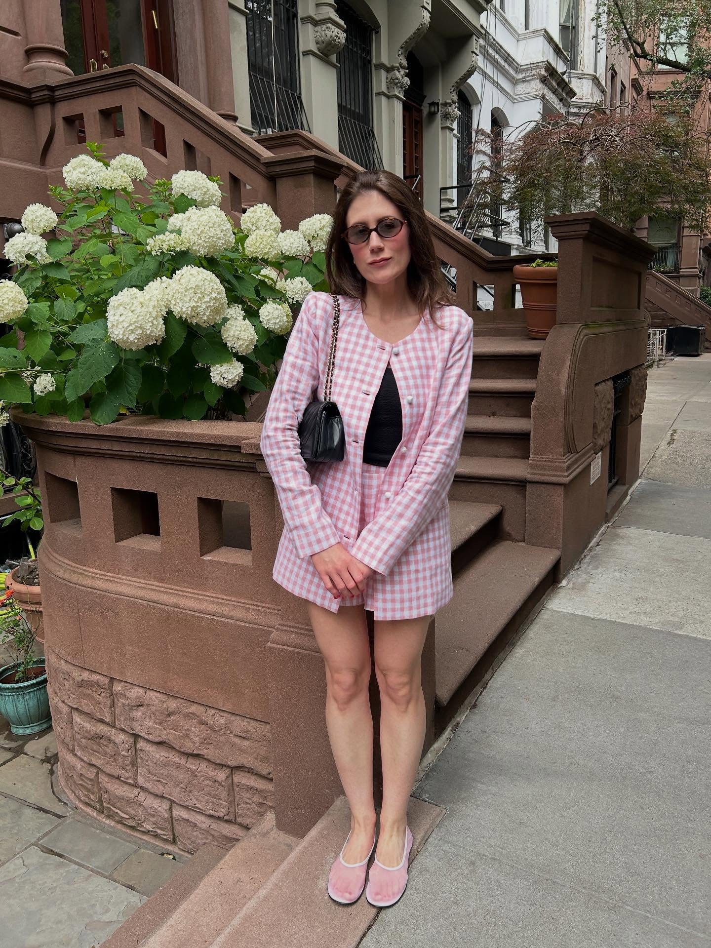Ally wears a pink check print jacket and shorts with mesh flats and a black top.