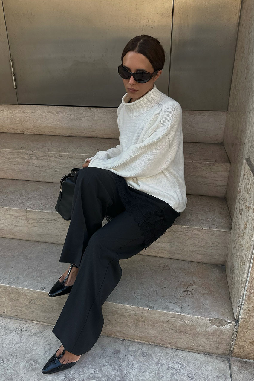 A woman wearing a cream sweater, black pants, and black pointed-toe heels