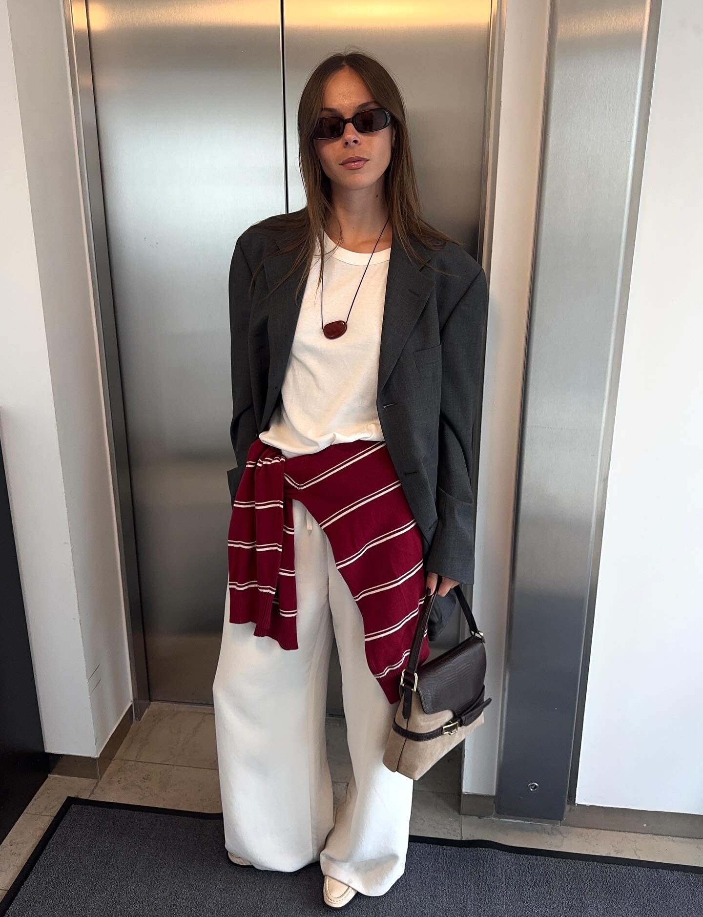 Swedish fashion creative Johanna Lager poses in a black rectangular sunglasses, an oversize gray blazer jacket, a white T-shirt, a cord necklace with a red pendant, a striped red sweater tied around the waist, white wide-leg jeans, an ATP Atelier shoudler bag, and cream loafers