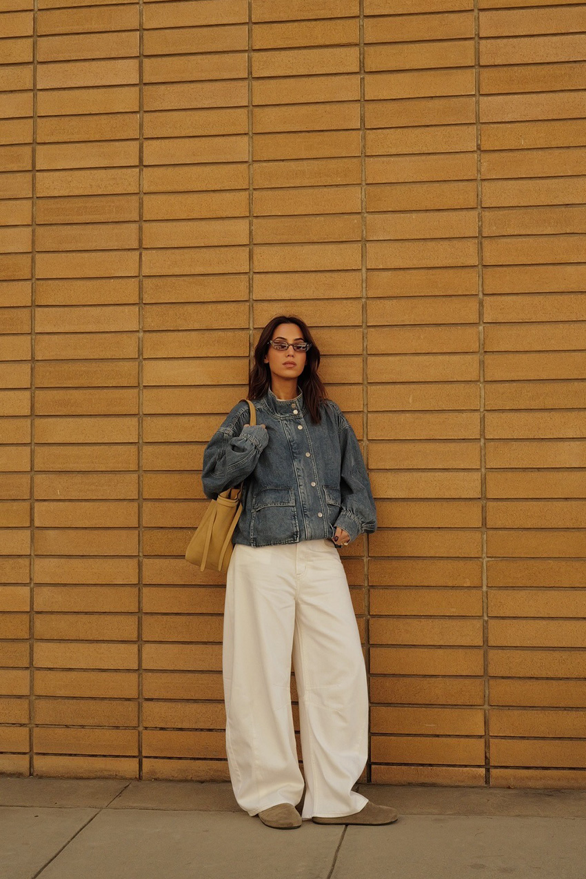 How to style white jeans for fall, shown in a photo of a woman standing outside against a yellow brick wall, wearing brown sunglasses, an oversize denim funnel neck jacket, a white t-shirt, white wide-leg jeans, suede Birkenstocks, and an oversize brown tote bag
