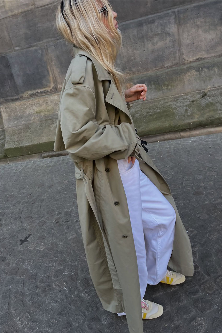 A woman wearing a tan trench coat, white jeans, and yellow sneakers