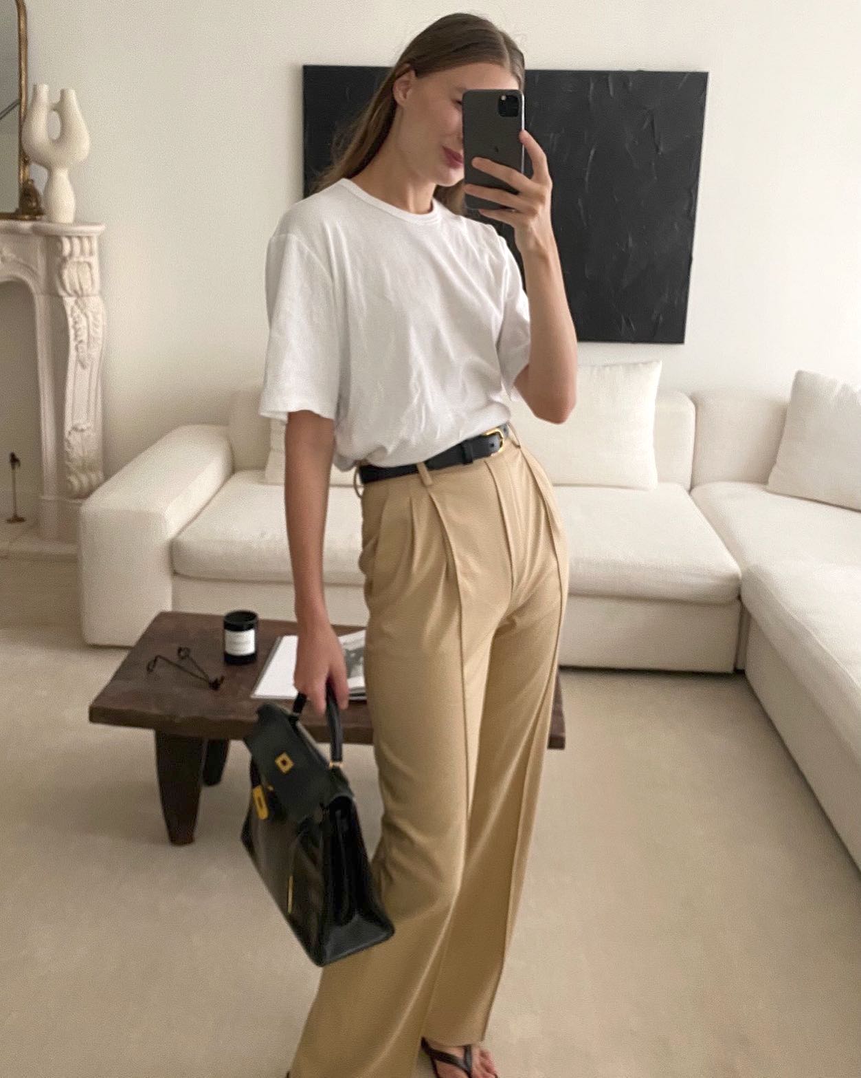 New York City fashion influencer Christie Tyler poses for a mirror selfie in her chic livingroom wearing a white t-shirt, black belt, vintage Hermes bag, high-waisted pleated tan pants, and flat black flip-flop sandals