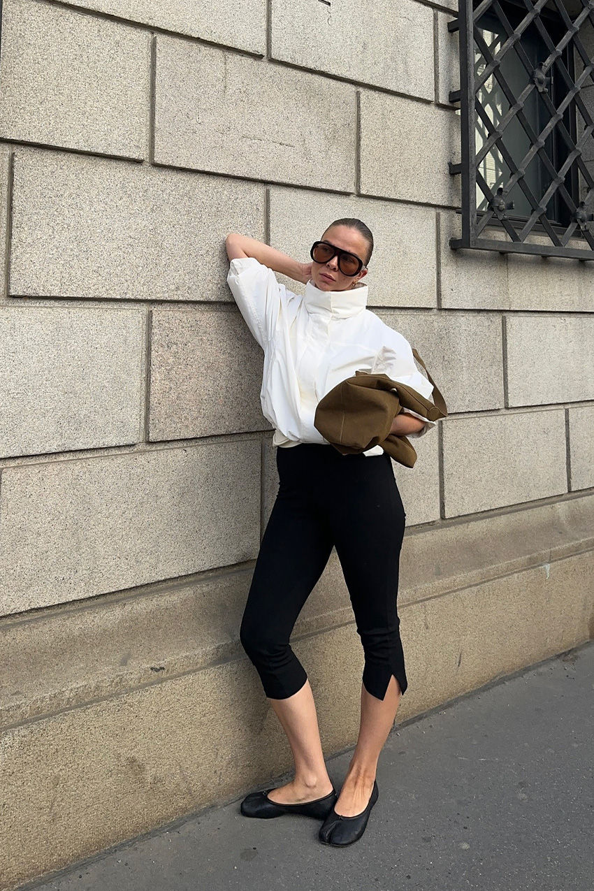 A windbreaker and plain leggings outfit idea is shown in a photo of a woman standing outside on a sidewalk, leaning against a gray wall, wearing oversized tinted aviator sunglasses with a white anorak jacket with cropped capri leggings, a suede hobo bag, and black Maison Marigela Tabi ballet flats