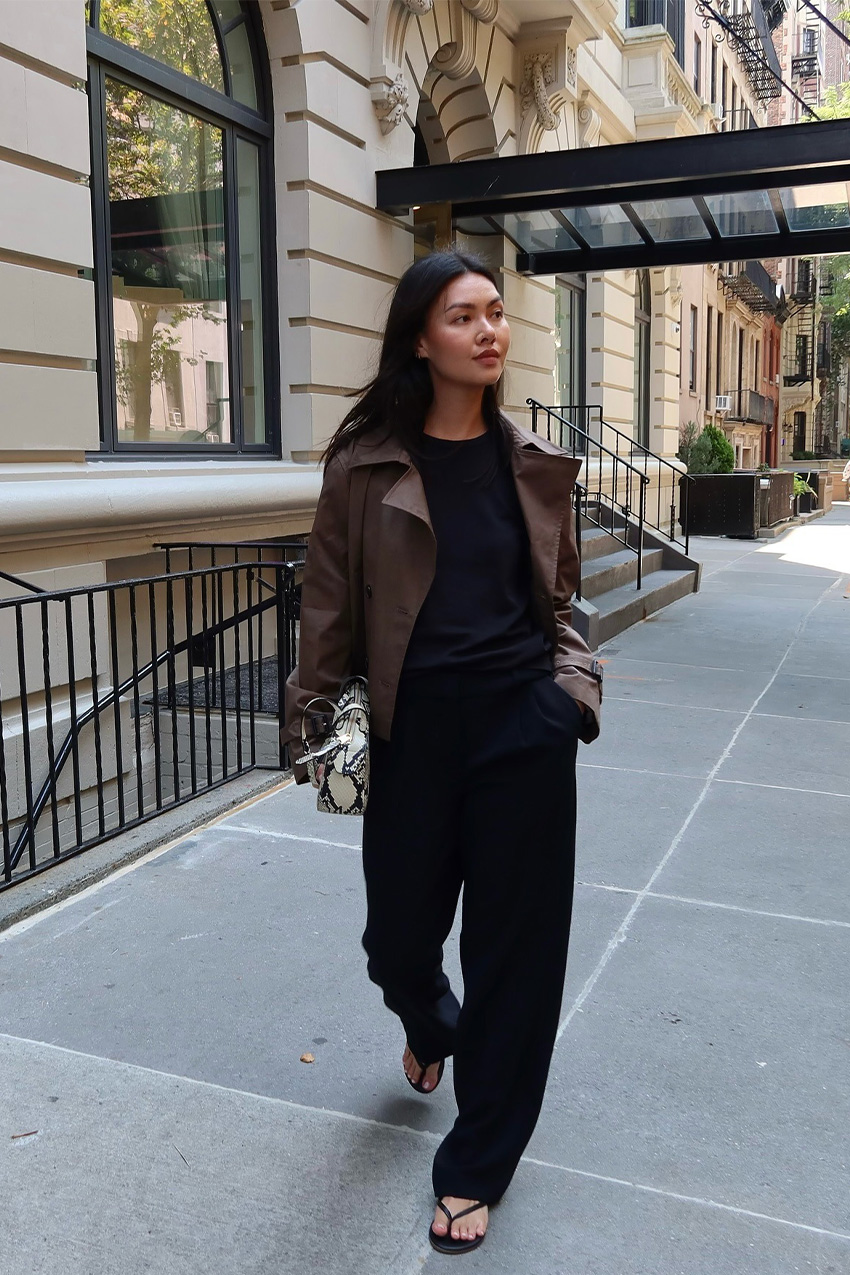 a flip-flop outfit for fall, shown in a photo of a woman walking down the street in New York wearing a cropped chocolate brown trench coat over a black crewneck sweater styled with black trousers, a gray and white snakeskin east-to-west bag, and black thong sandals