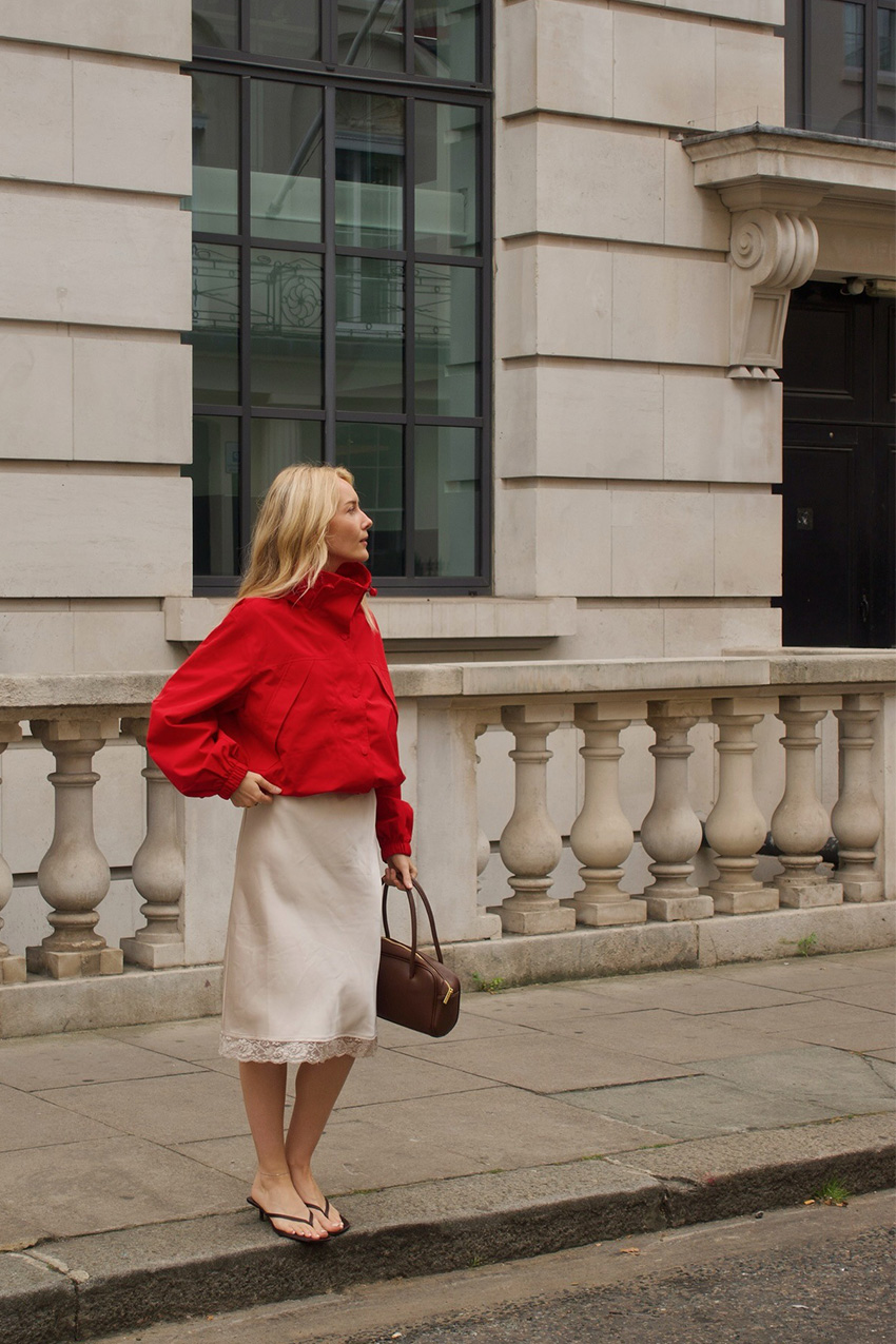 a flip-flop outfit for fall, shown in a photo of a woman standing on the sidewalk in London wearing a red windbreaker jacket with a white slip skirt, a brown bowler bag, and black heeled flip-flop sandals