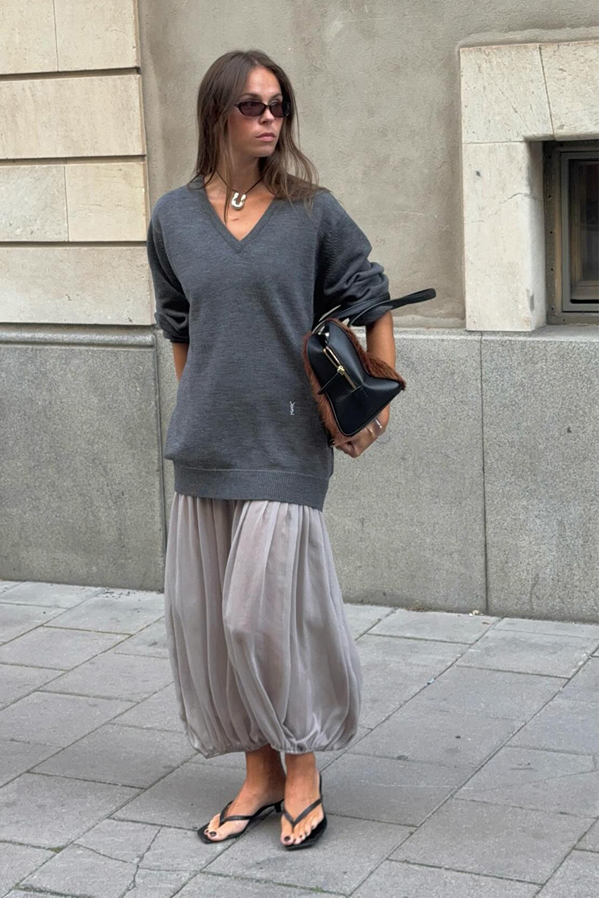 a flip-flop outfit for fall, shown in a photo of a woman standing on the sidewalk wearing black sunglasses, a black cord necklace, a gray oversized V-neck sweater, gray sheer ballon pants, black heeled thong sandals, and a two-tone bowler bag