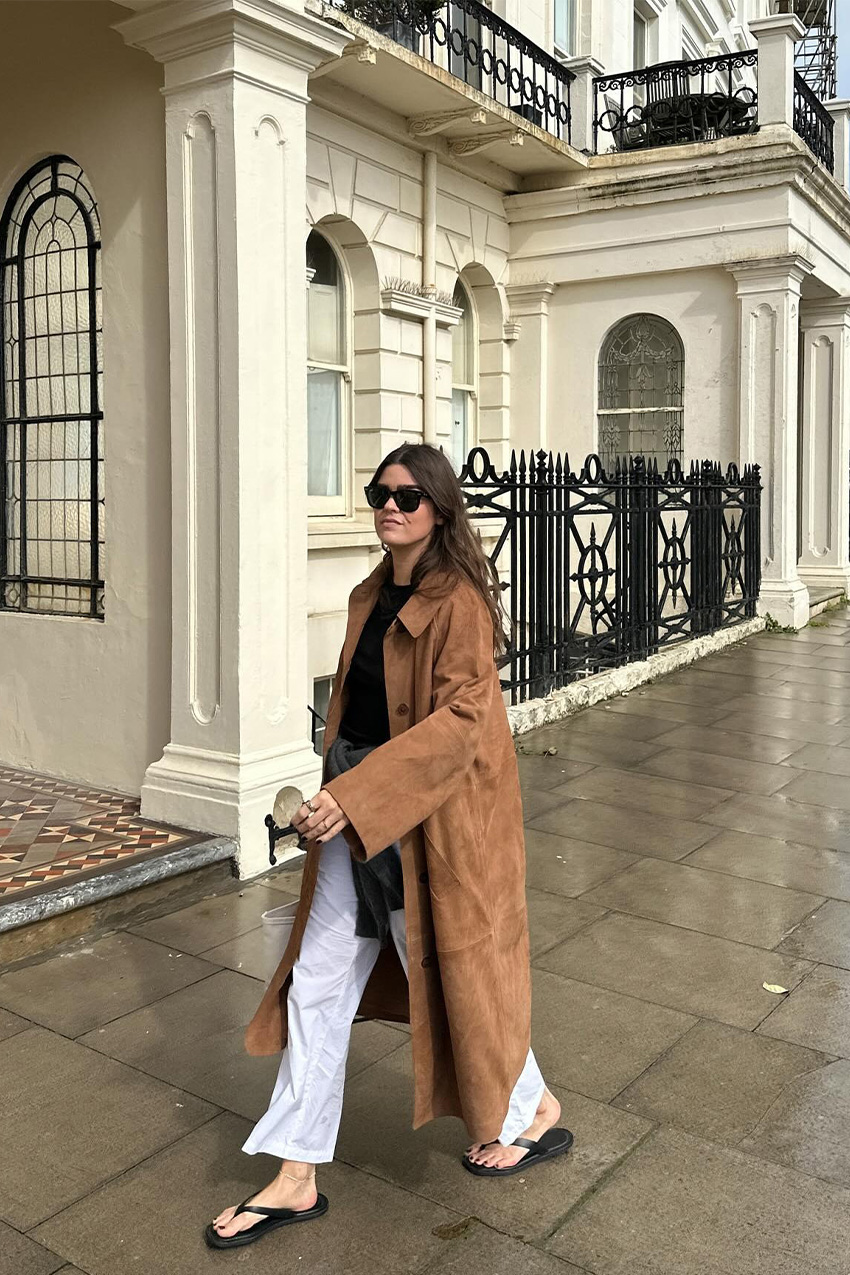 a flip-flop outfit for fall, shown in a photo of a woman walking down the sidewalk in London wearing a long tan suede coat styled over a black crewneck sweater with white trousers and a black cardigan tied around the waist, worn with black flip-flop sandals, black sunglasses, and a white tote bag