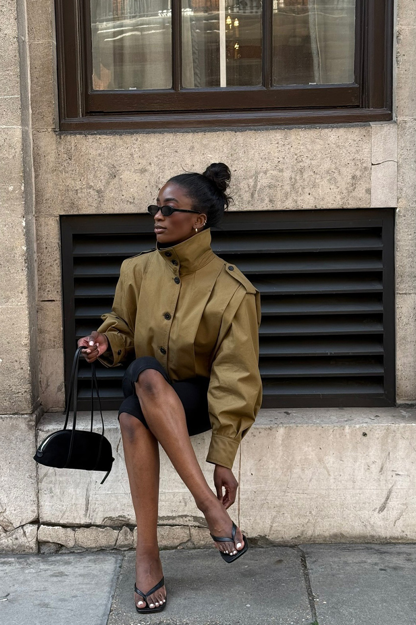 a windbreaker and plain leggings outfit idea is shown in a photo of a woman sitting on a ledge outside on the sidewalk wearing black sunglasses, stacked silver ear cuff, a tan parka jacket, black capri leggings, black heeled thong sandals, and a black shoulder bag