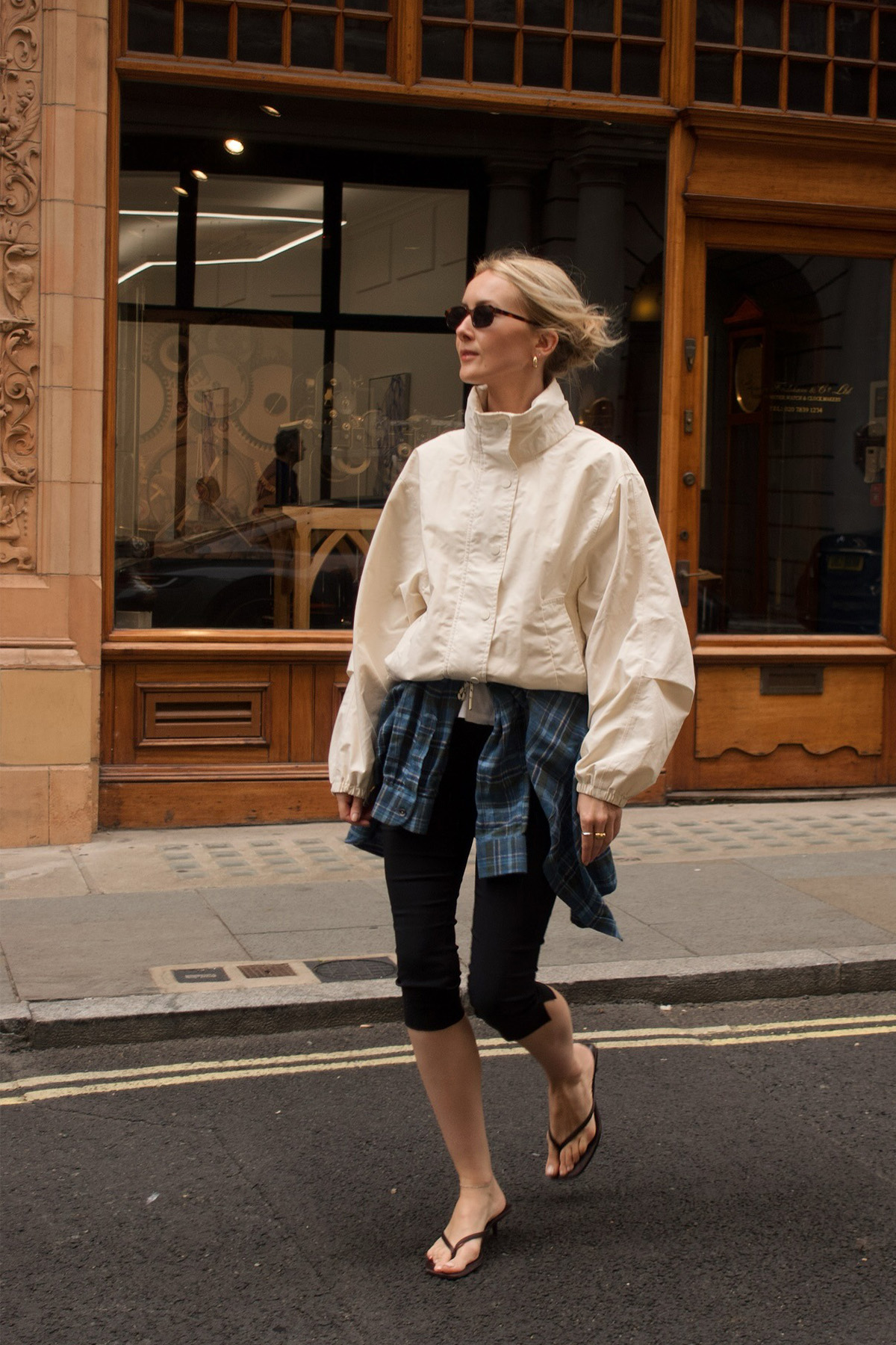 a windbreaker and plain leggings outfit idea is shown in a photo of a woman crossing the street wearing skinny sunglasses, with a white parka jacket, black capri leggings, a blue plaid fleece button-down shirt tied around the waist, and black flip-flops