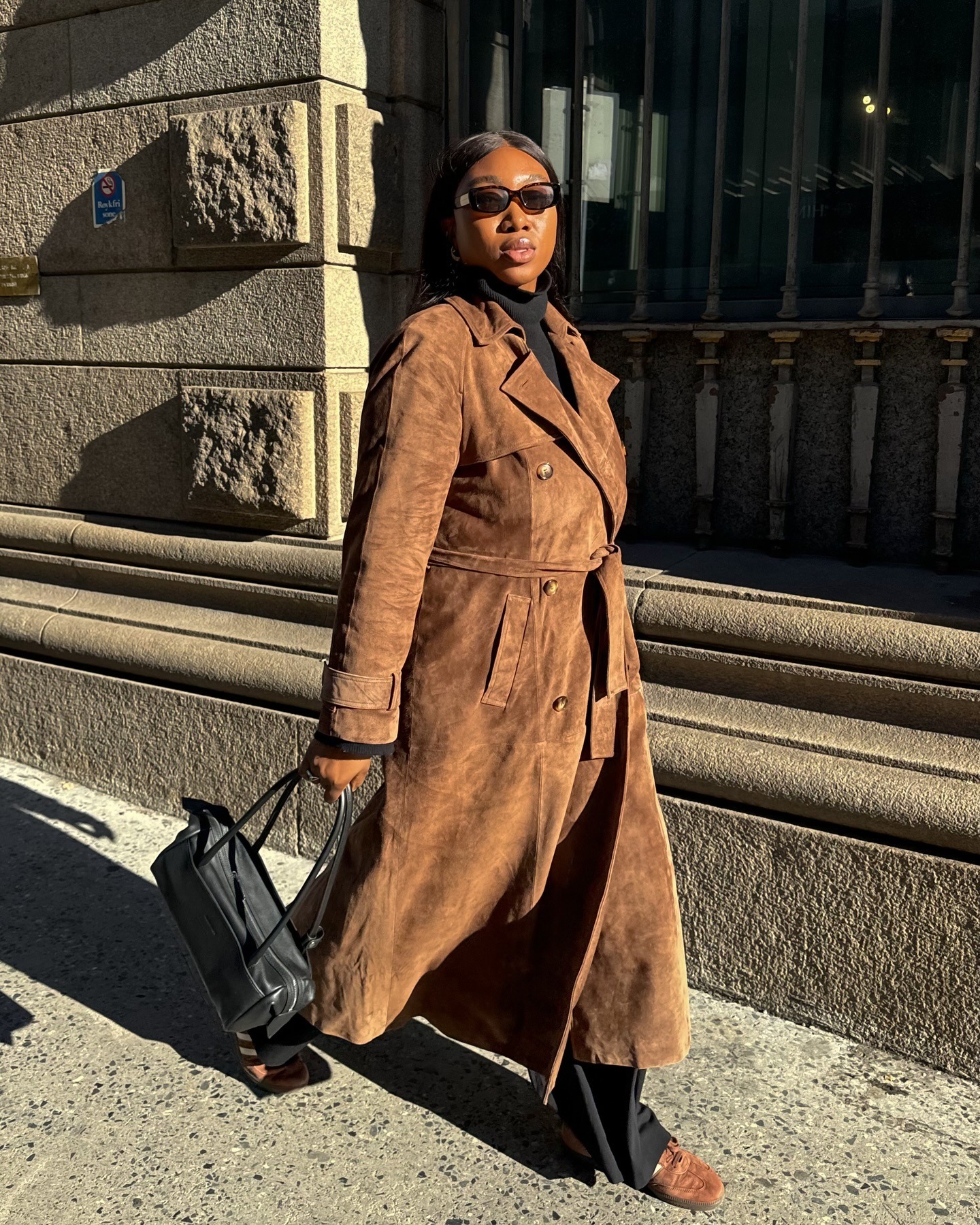 Influencer Nnenna wears a suede long coat, black East West bag, black slacks, and suede Adidas sneakers.