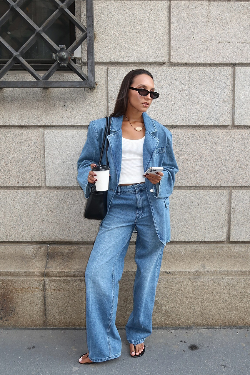 a flip-flop outfit for fall, shown in a photo of a woman standing on a sidewalk against a brick wall wearing black sunglasses with a denim blazer, silver bean cord necklace, a white tank top, light-wash wide-leg jeans, black flip-flops, and a black hobo tote bag