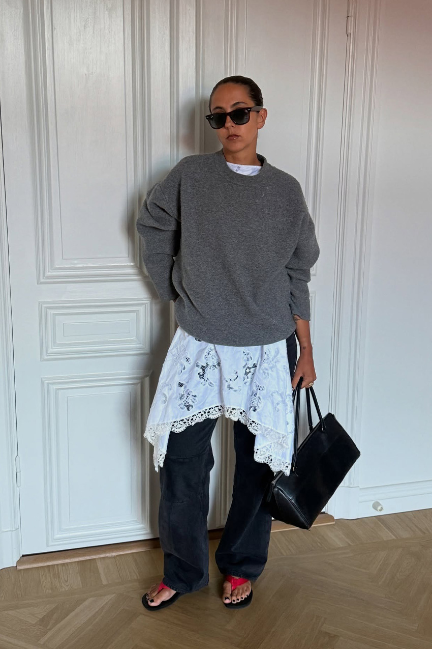 a flip-flop outfit for fall, shown in a photo of a woman standing inside an apartment with white walls and wool floors, wearing black sunglasses with a gray oversized sweater styled over a white lace dress that's layered over baggy black jeans and styled with a black handbag and black and red flip-flops