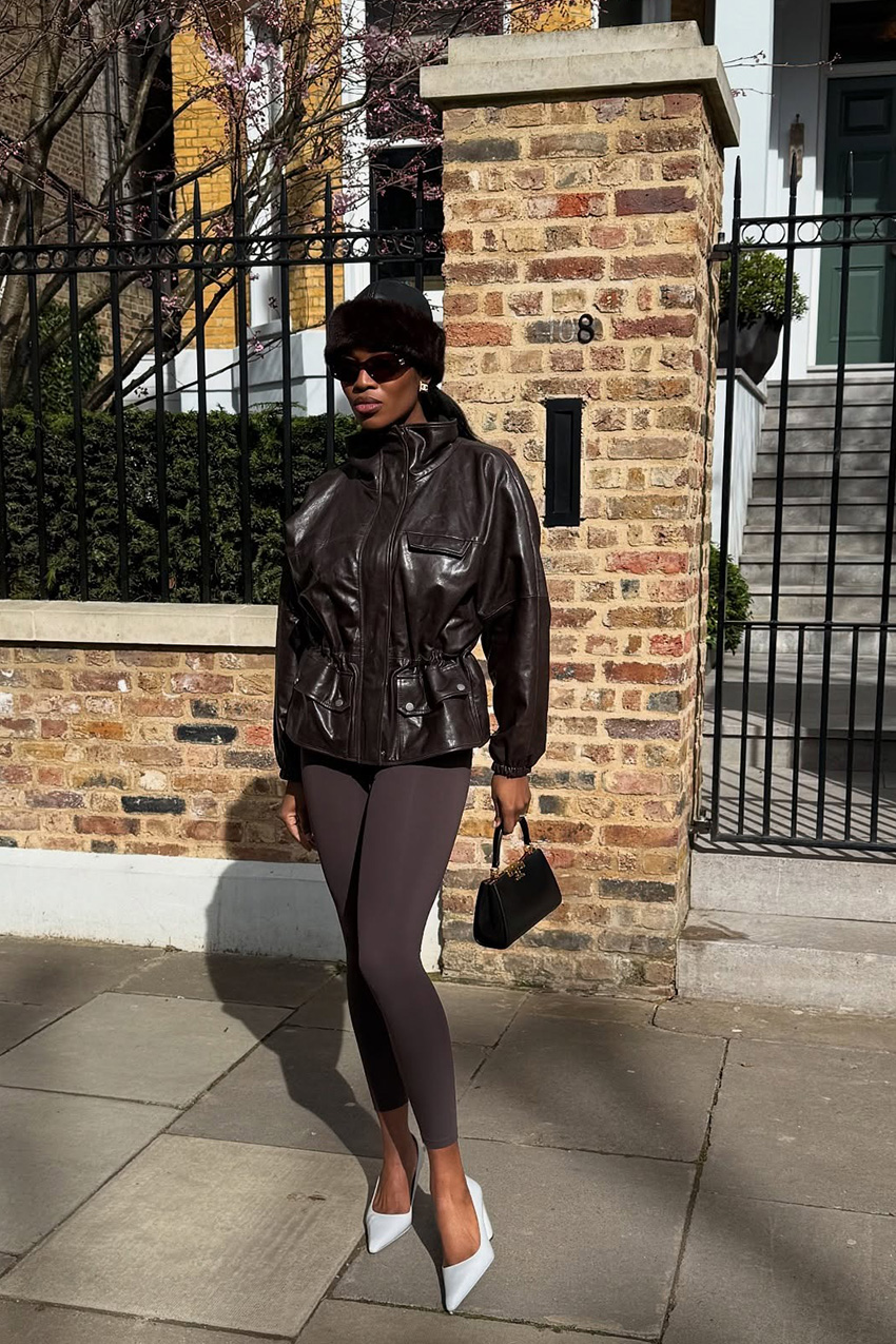 A windbreaker and plain leggings outfit idea is shown in a photo of a woman standing outside on the sidewalk wearing a fur hat with black sunglasses, gold clip-on earrings, a faux leather brown parka jacket, brown plain leggings, white pointed pumps, and a small black Tory Burch handbag