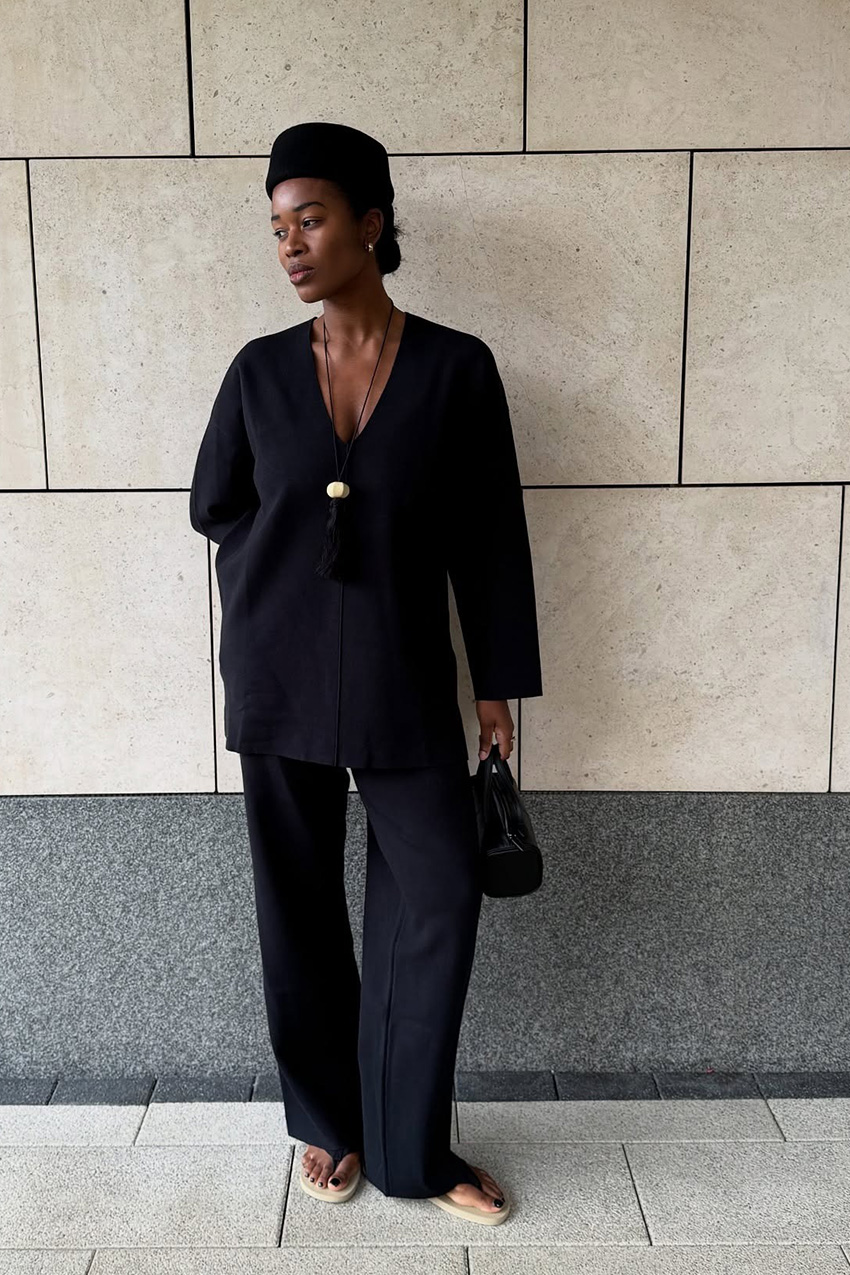 a flip-flop outfit for fall, shown in a photo of a woman standing outside, leaning against a gray wall, wearing a black box hat with a black V-neck sweater, a long tassel necklace with a creme stone, black tailored trousers, a black barrel handbag, and tan and black flat thong sandals