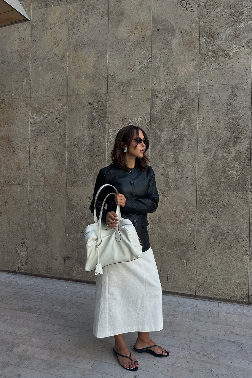a flip-flop outfit for fall, shown in a photo of a woman standing outside against a concrete backdrop wearing black sunglasses, silver earrings, a black leather blazer, a white pencil skirt, a white bowler bag, and black flip-flops