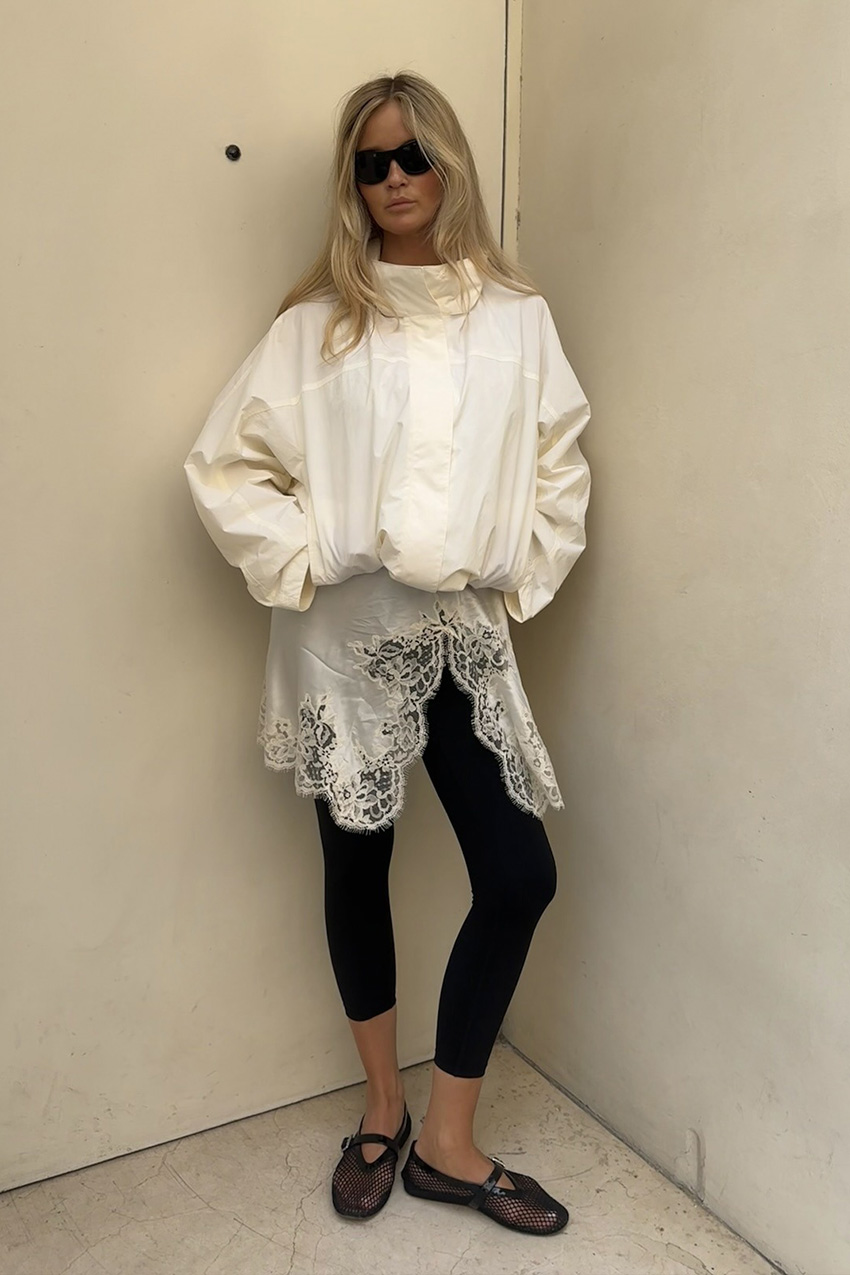 a windbreaker and plain leggings outfit idea is shown in a photo of a woman standing against a white wall wearing an oversized white parka jacket over a white lace satin slip dress styled with plain black leggings, black mesh ballet flats, and black sunglasses