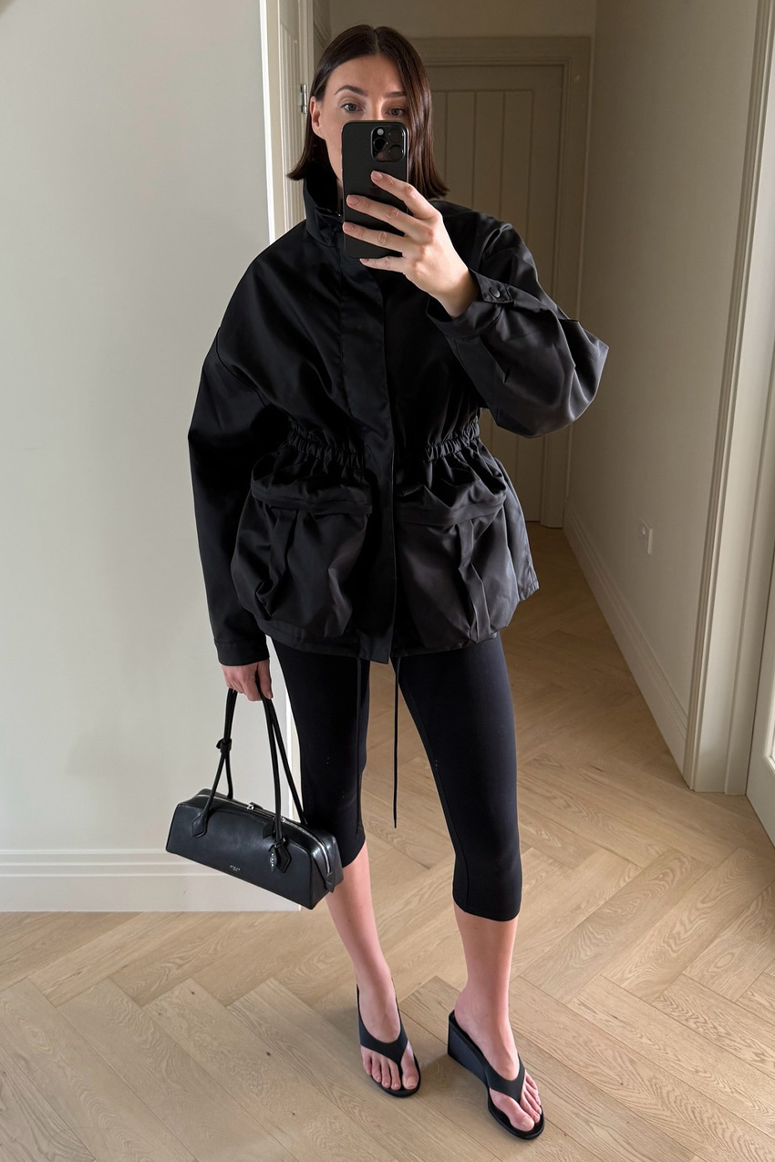 A windbreaker and plain leggings outfit idea is shown in a photo of a woman taking a mirror selfie in a minimal hallway of their home wearing a large black parka with black cropped leggings, black thong heeled sandals, and a Le Tekel Alaia bag