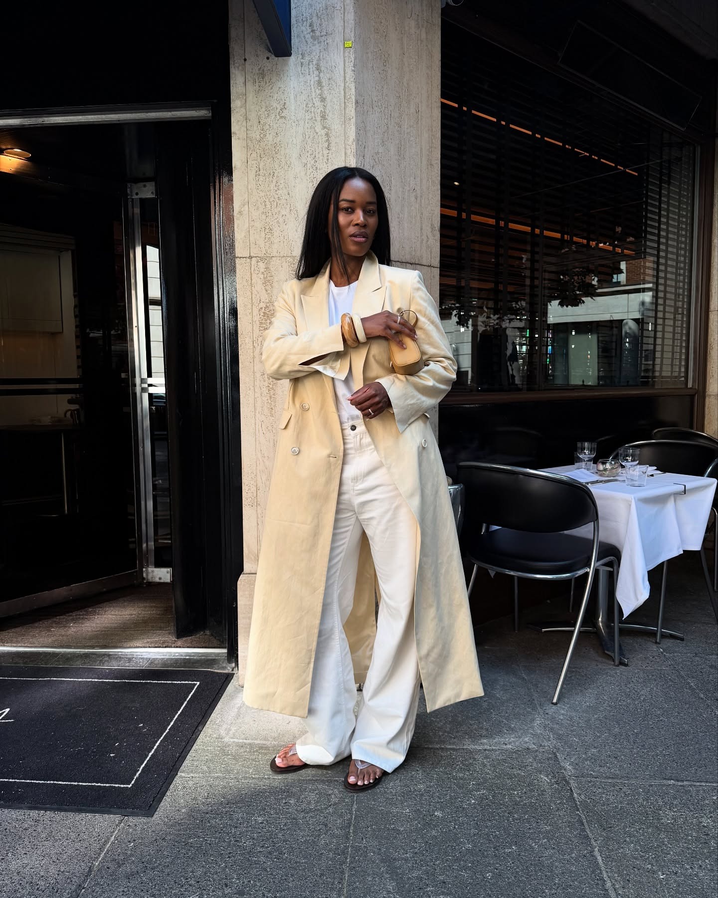 European style influencer Marilyn Nwawulor-Kazemaks wearing chic fall outfit with white jeans, a pale yellow maxi coat, white T-shirt, bangles, mini yellow bag, and clear thong flip-flop sandals