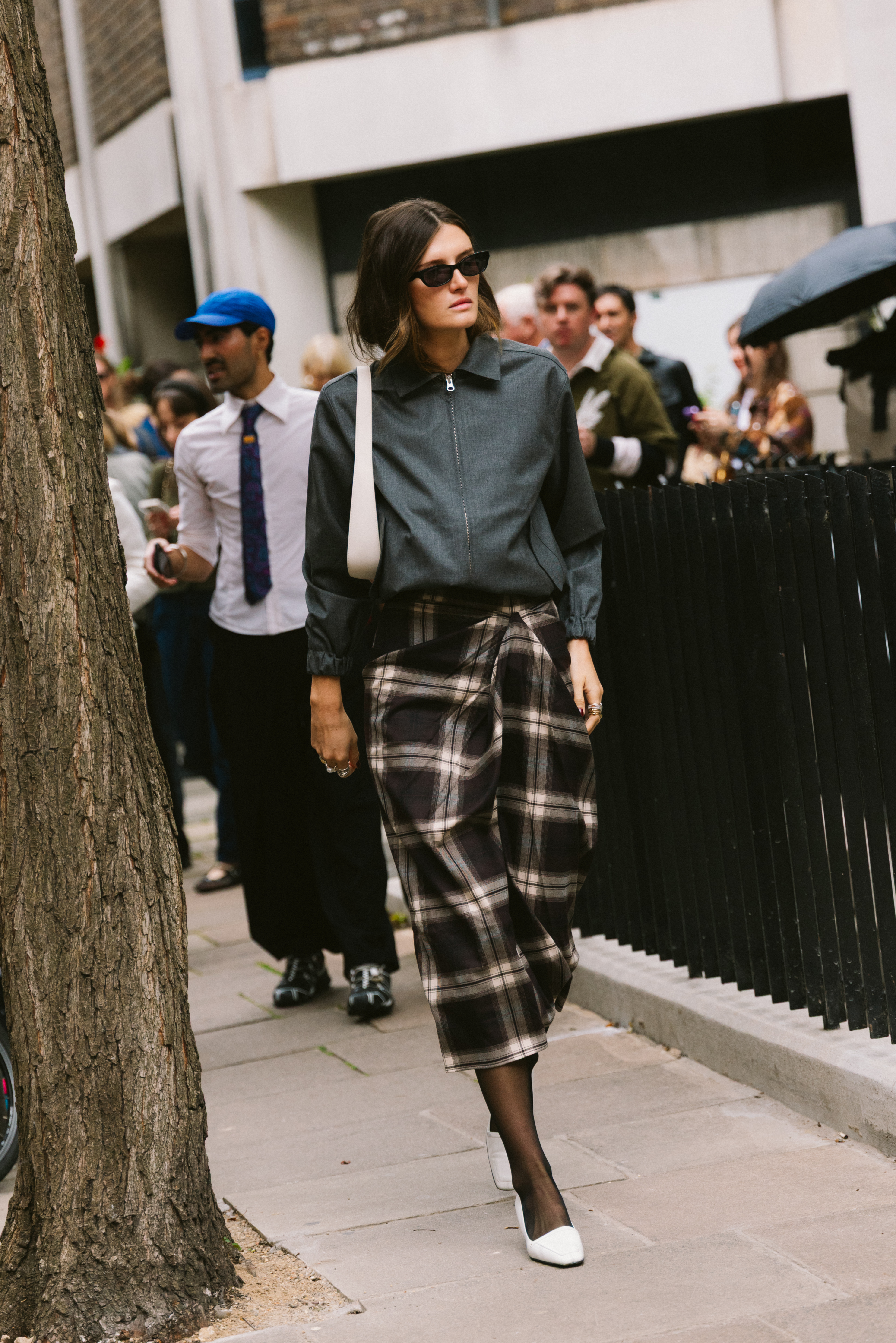 A woman wearing a gray jacket, plaid skirt, and white heels