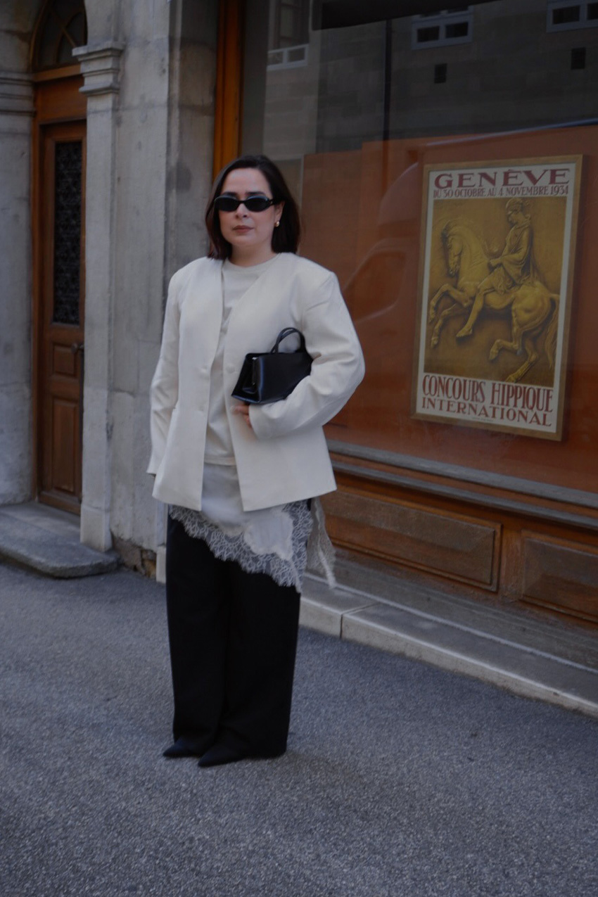 A sweater styling trend is shown in a photo of a woman's outfit, where she's standing outside in front of a storefront wearing a white blazer over a white cashmere t-shirt layered over a white slip dress with black wide-leg jeans, black pointed heels, and a black frame bag