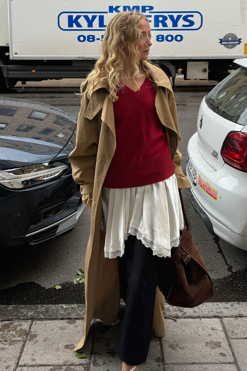 A sweater styling trend is shown in a photo of a woman's outfit where she's standing on the sidewalk in front of parked cars wearing a long tan trench coat on top of red v-neck sweater that's layered over a white slip dress and black jeans styled with white cutout heels and a chocolate brown suede bowler bag