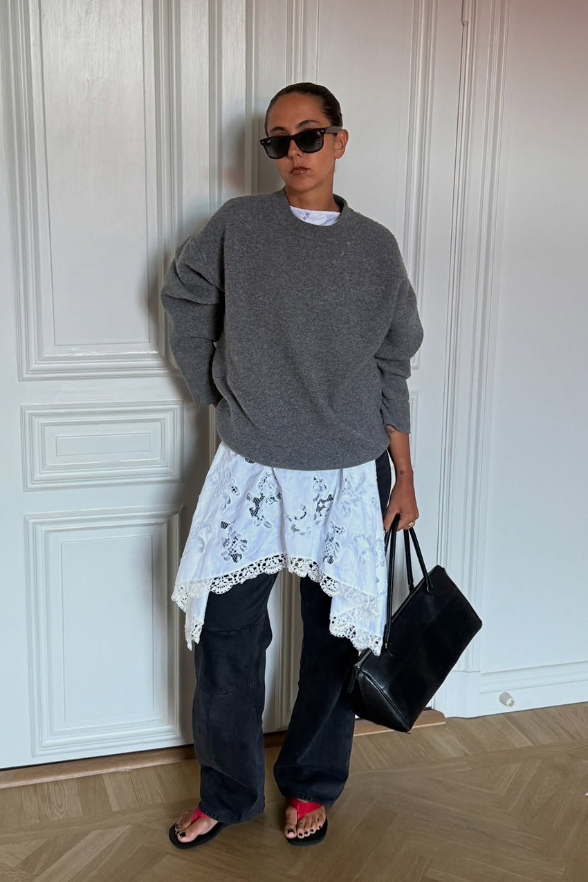 A sweater styling trend is shown in a photo of a woman's outfit, where she's standing inside her home against a white wall, wearing an oversize gray crewneck sweater over a white lace slip dress styled over black baggy jeans with red and black flip flops, black sunglasses, and a black handbag