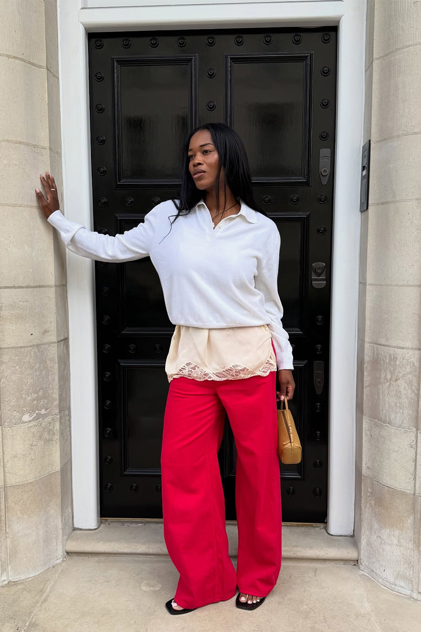 A sweater styling trend is shown in a photo of a woman's outfit where she's standing in a doorway outside wearing a white polo sweater over a cream lace slip dress, and red jeans worn with black sandals and a brown barrel bag