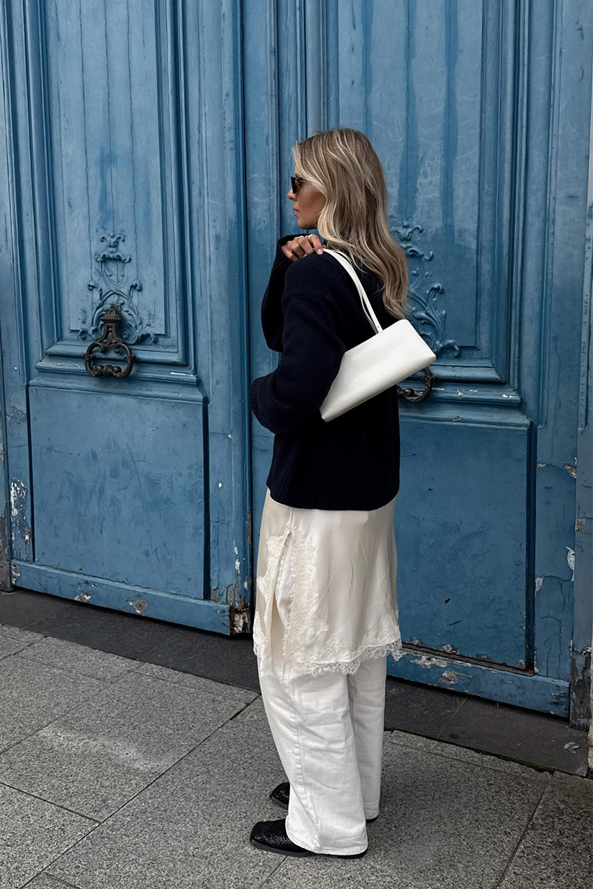 A sweater styling trend is shown in a photo of a woman's outfit, where she's standing outside in front of a blue doorway in Paris wearing a navy blue turtleneck sweater over a white slip dress and matching white wide leg jeans styled with black ankle boots and a white shoulder bag