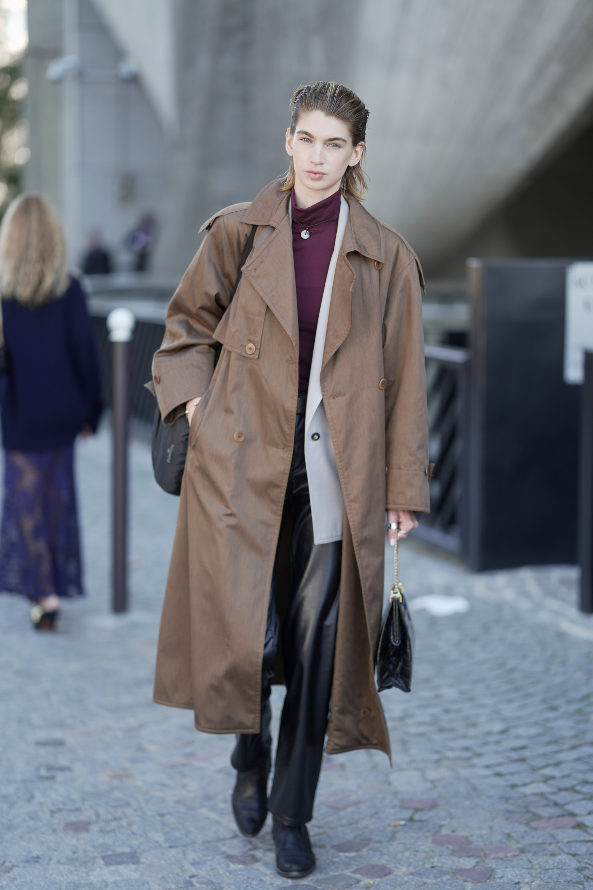 A woman wears a brown trench coat, burgundy turtleneck, black leather pants, and black ankle boots.