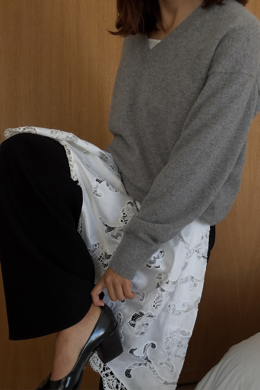 A sweater styling trend is shown in a close photo of a woman's outfit, where she's standing in a hotel room wearing a gray sweater over a white lace dress worn over black jeans styled with black kitten heel pumps