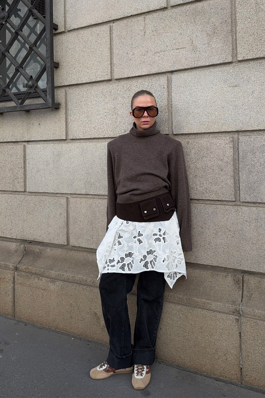 A sweater styling trend is shown in a photo of a woman's outfit, where she's standing outside on a sidewalk, leaning against a brick wall, wearing oversize two-tone aviator sunglasses with a brown turtleneck sweater that's layered over a white lace dress and baggy black jeans, styled with a belt bag and suede tan sneakers