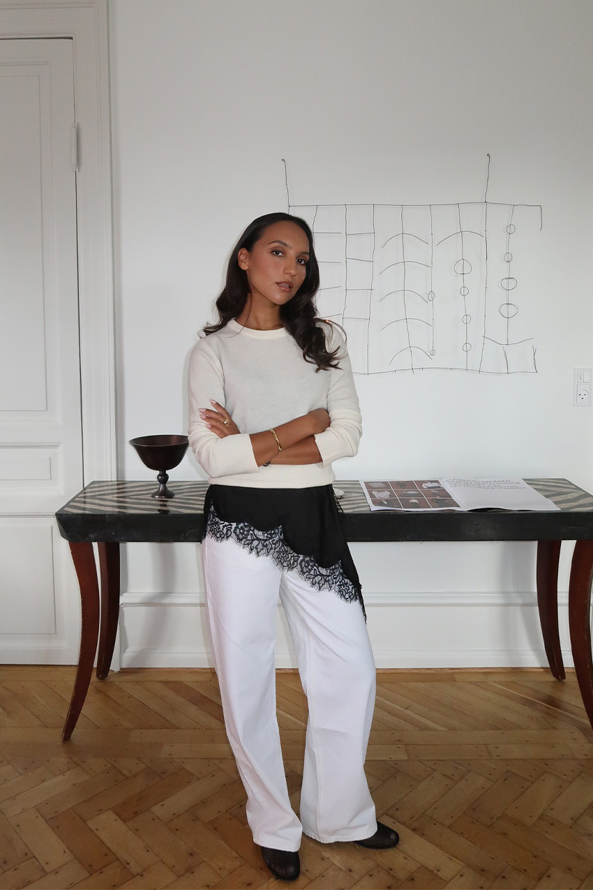 A sweater styling trend is shown in a photo of a woman's outfit, where she's standing inside her home in a hallway with a table and art behind her, wearing a white crewneck sweater over a black slip dress layered on top of white baggy jeans with black flats