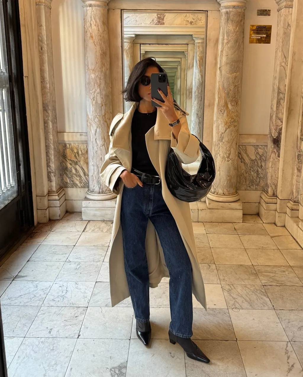 Shay wears a trench coat, black shirt, blue jeans, black belt, black kitten-heeled boots and a black khaite bag.