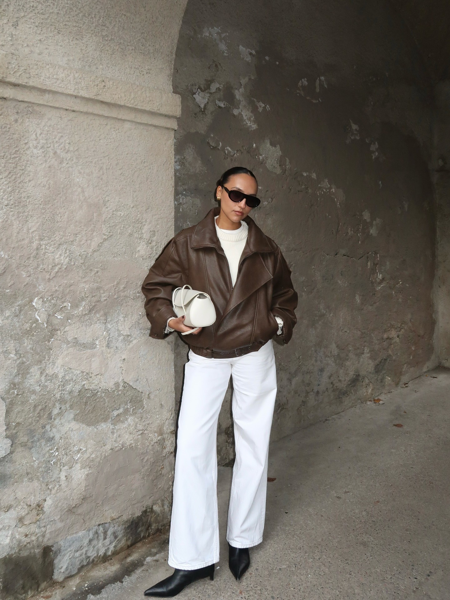 Ingrid wears a brown leather jacket, white sweater, white bag, white leather pants, and black kitten heel boots.