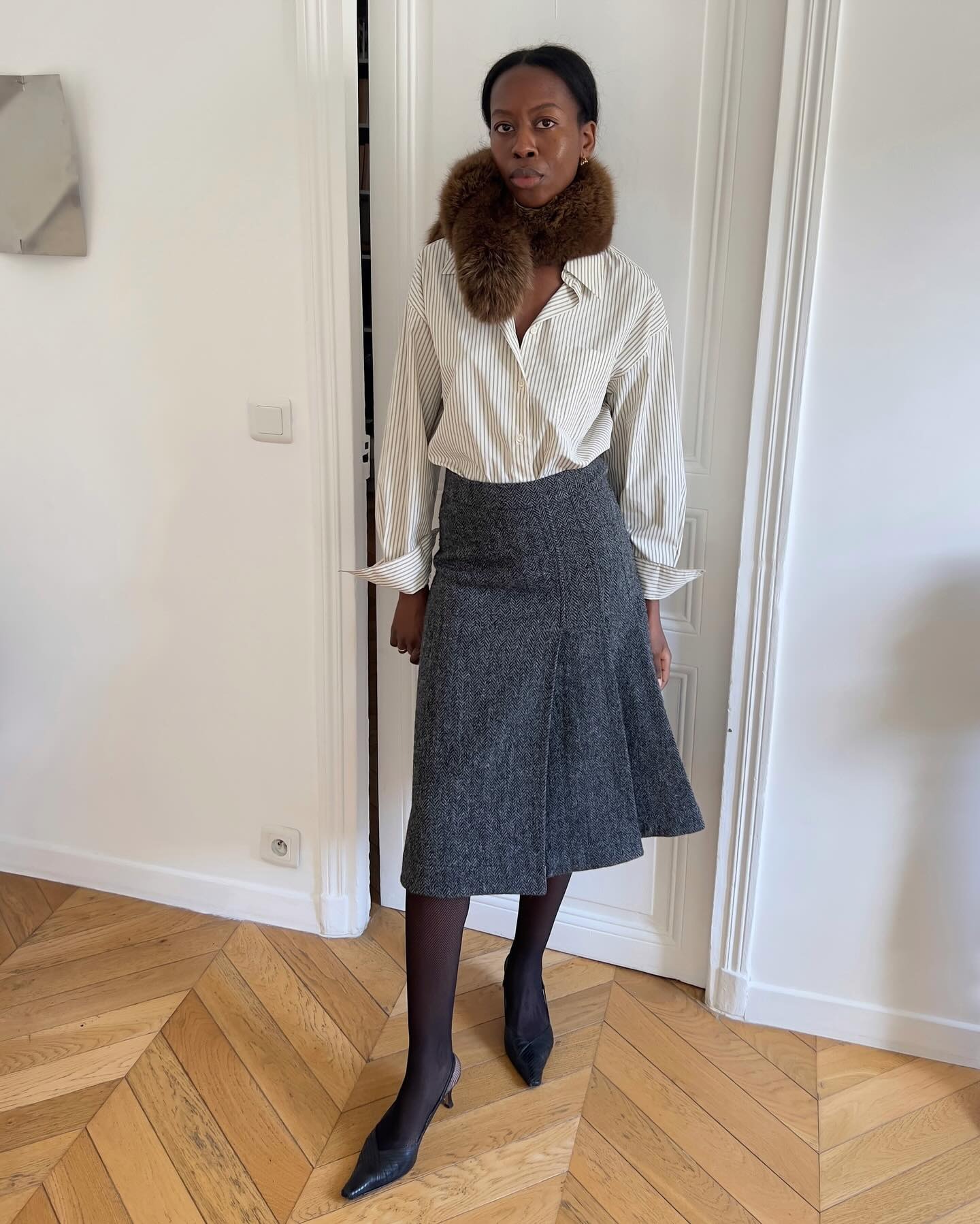 Sylvie Mus wearing a skirt outfit idea for 2026, including a gray pleated midi skirt with a button-down shirt and fur scarf.