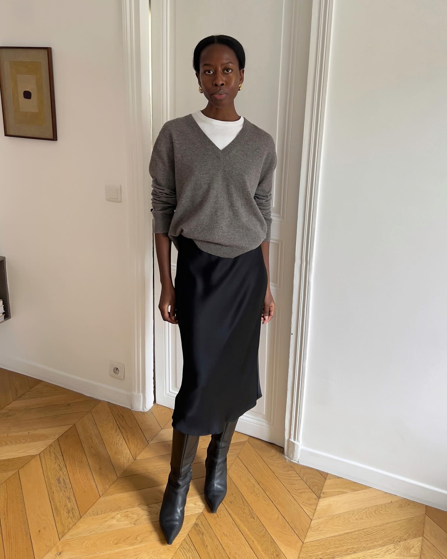 Influencer Sylvie Mus wears a silk black skirt, knee-high leather boots, a white tee, and a high-V neck heather grey sweater.