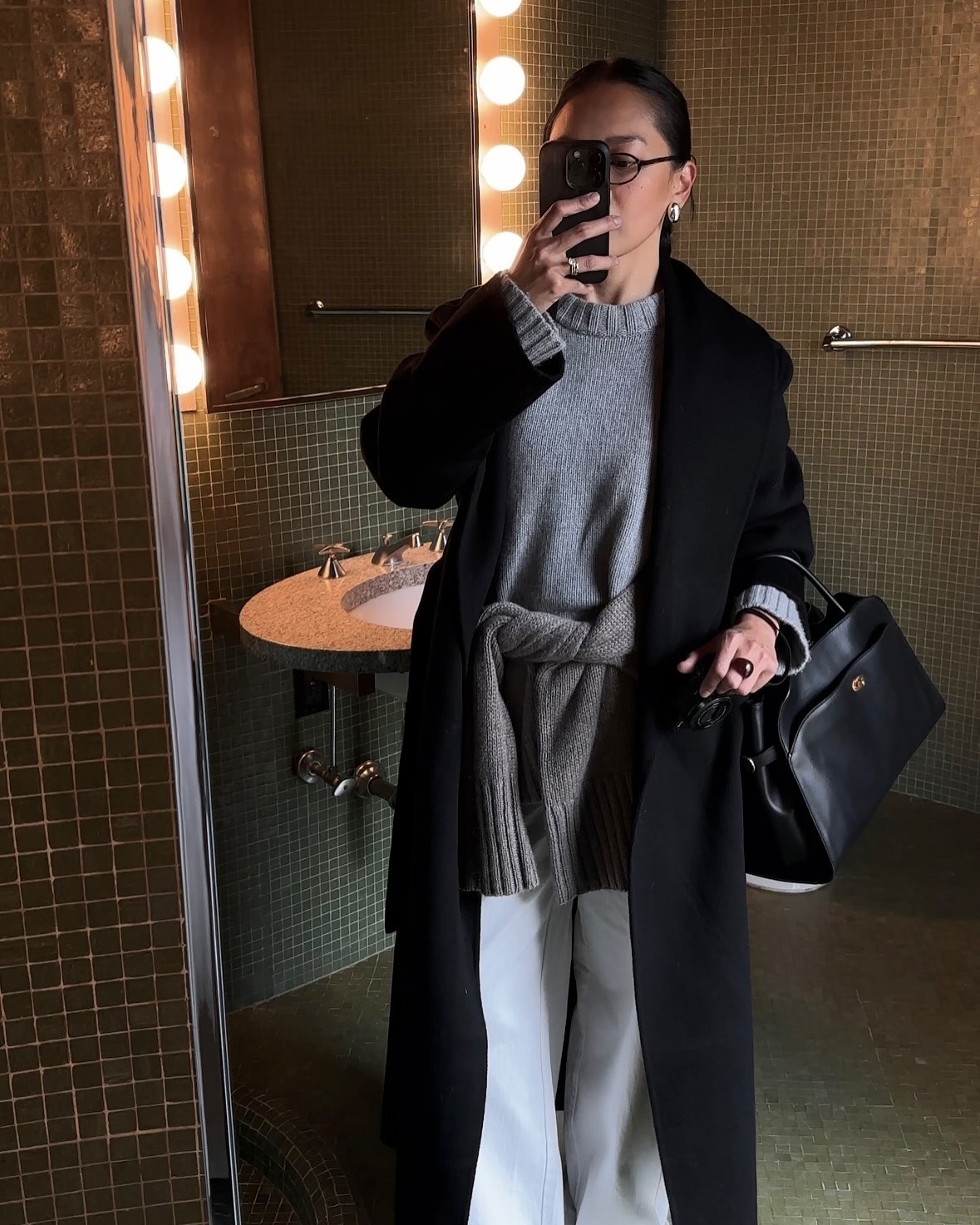 Influencer Dawn Tan wears white jeans, a grey sweater with a second tied around her waist, a black overcoat, glasses, and leather handbag.