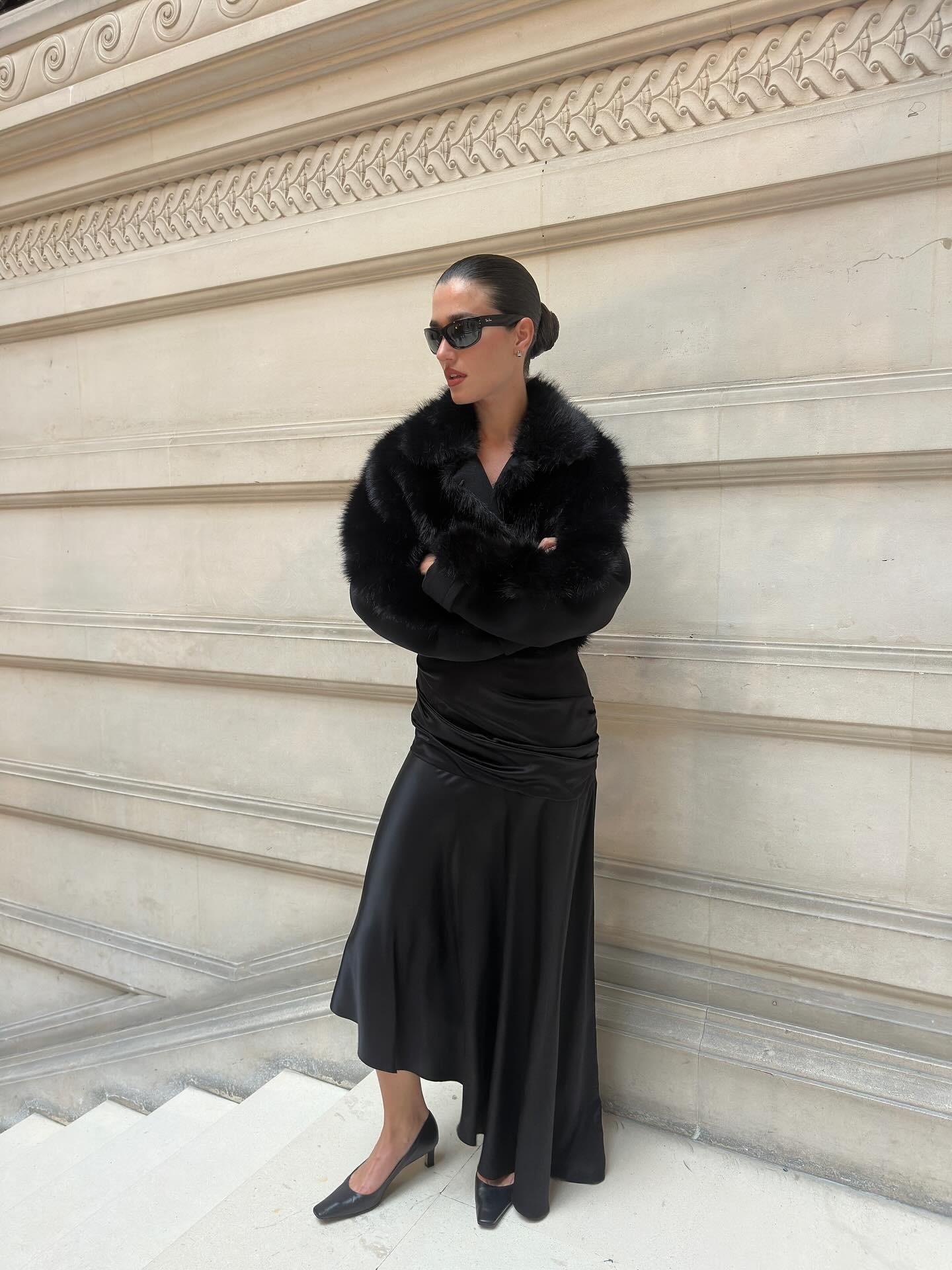 A woman wears a black fur coat, black dress, and black pumps.