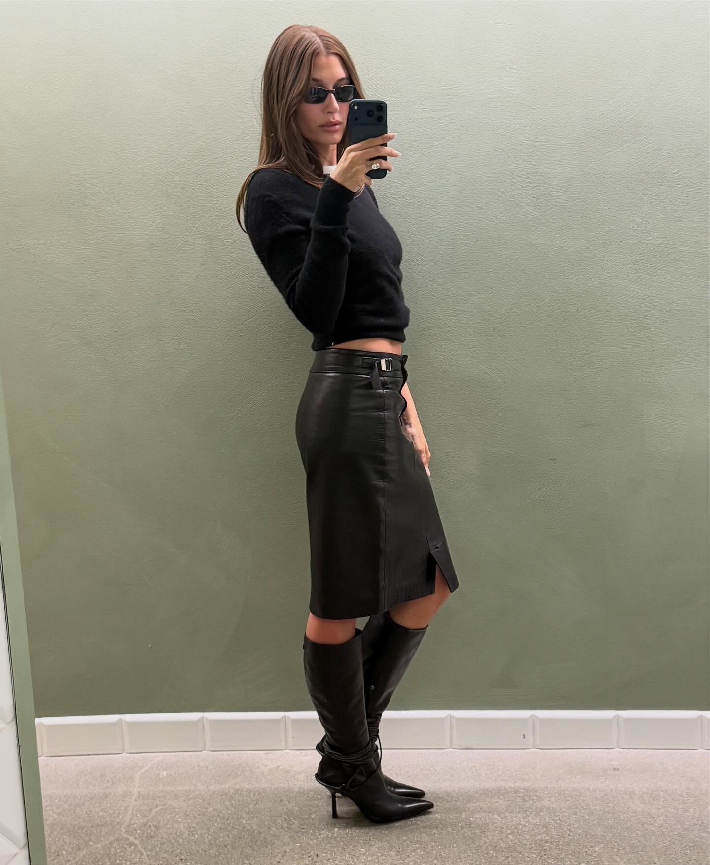 Hailey Bieber wearing a skirt outfit idea for 2026, including a black sweater with a black leather pencil skirt and boots.