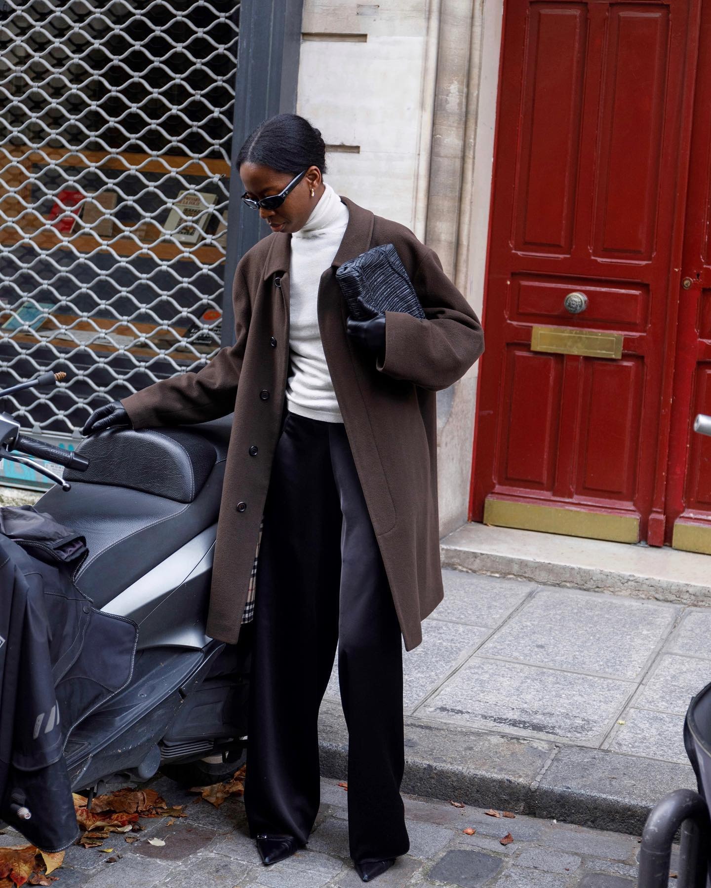 Style influencer Sylvie Mus poses next to a scooter in Paris wearing black oval sunglasses, a white turtleneck, brown Burberry coat, black leather clutch, black silk pants, and pointed-toe heels with a buckle detail