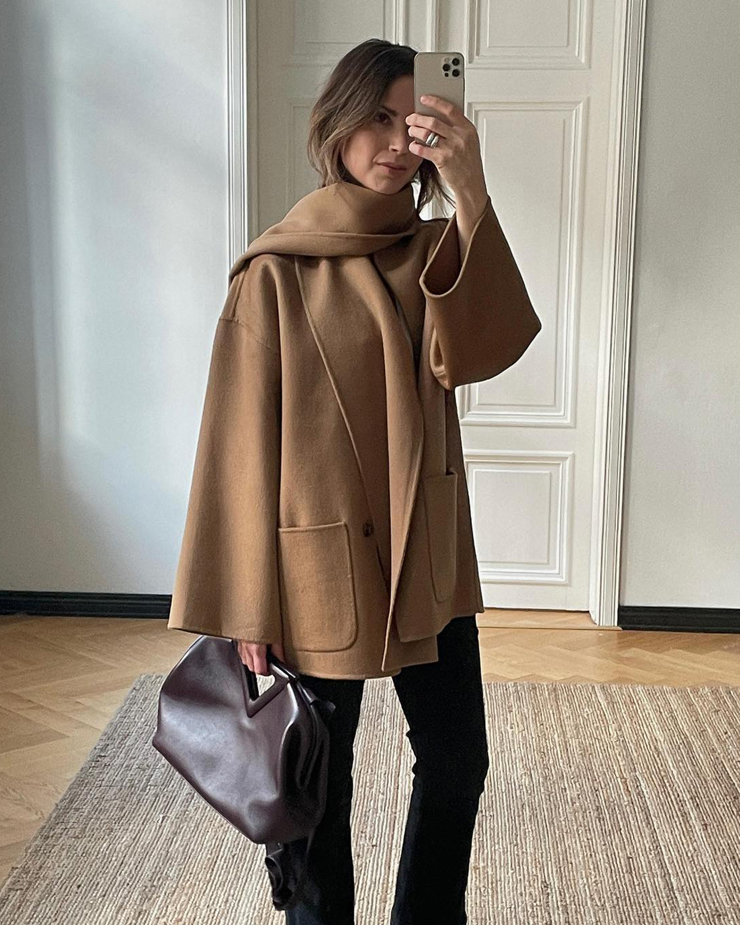style influencer Johanna Piispa poses for a mirror selfie wearing a camel scarf coat, burgundy Bottega Veneta bag, and black jeans