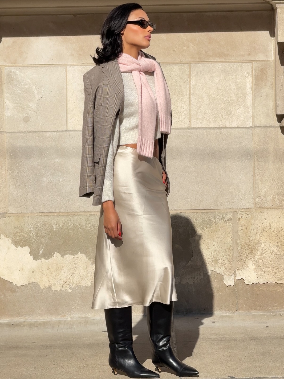 Woman wearing a white sweater, gold silk skirt, and black boots with a blazer over her shoulders and a pink sweater tied around her neck.