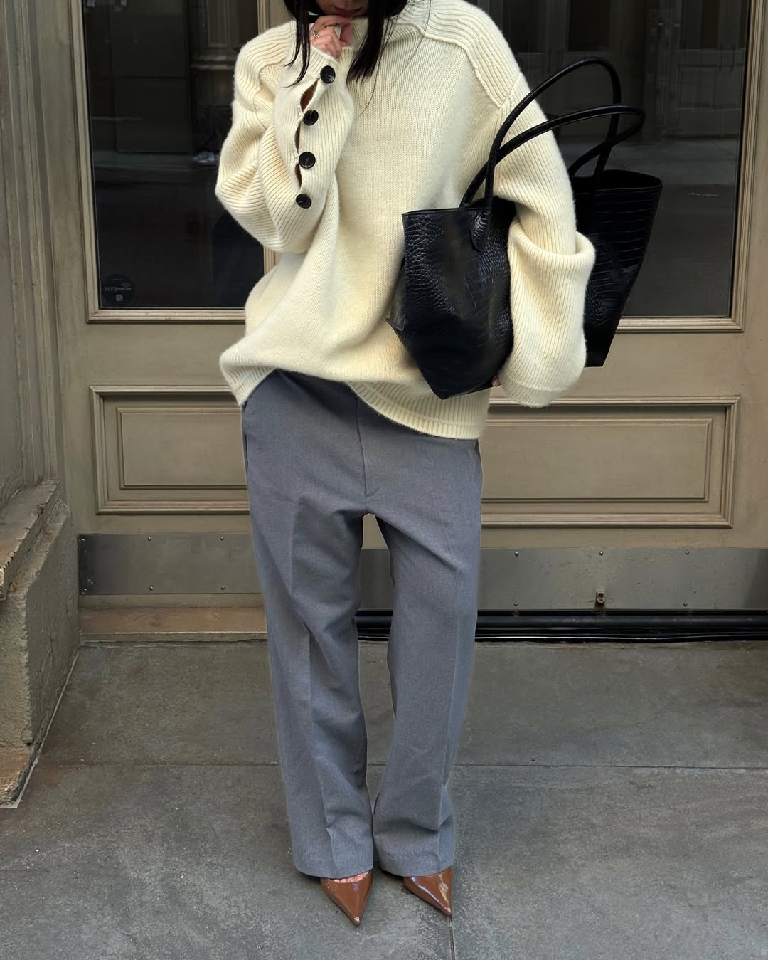 A woman wears a cream sweater, gray pants, and brown pumps with a black tote bag.