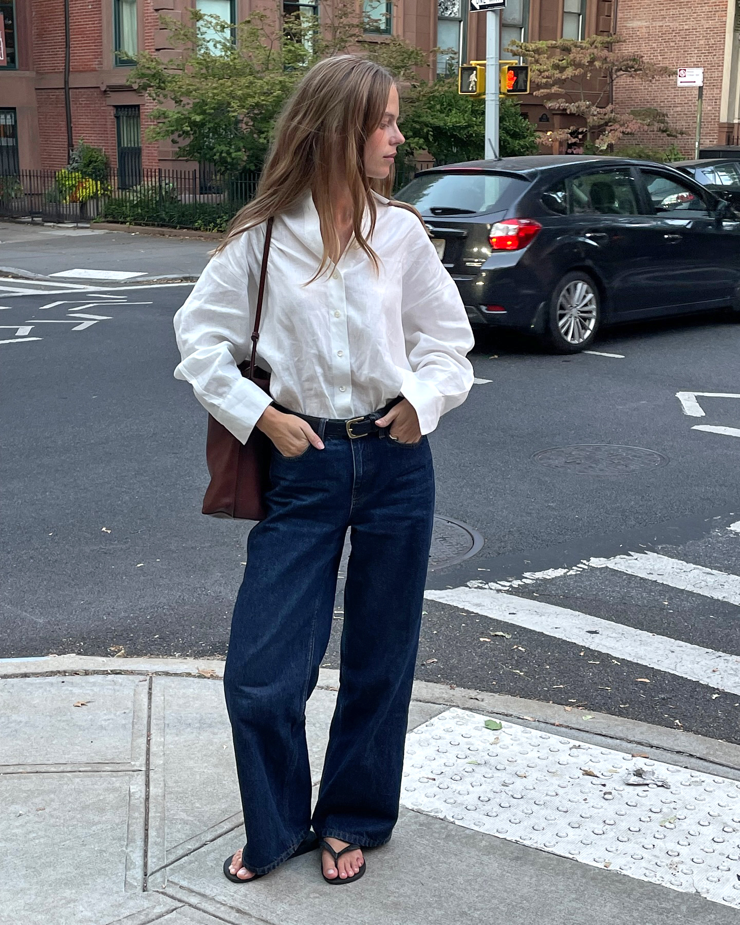 Danish fashion creative Clara Dyrhauge poses on a NYC street wearing a white linen button-down shirt, a brown tote bag, black belt, dark-wash baggy jeans, and black flip-flop sandals