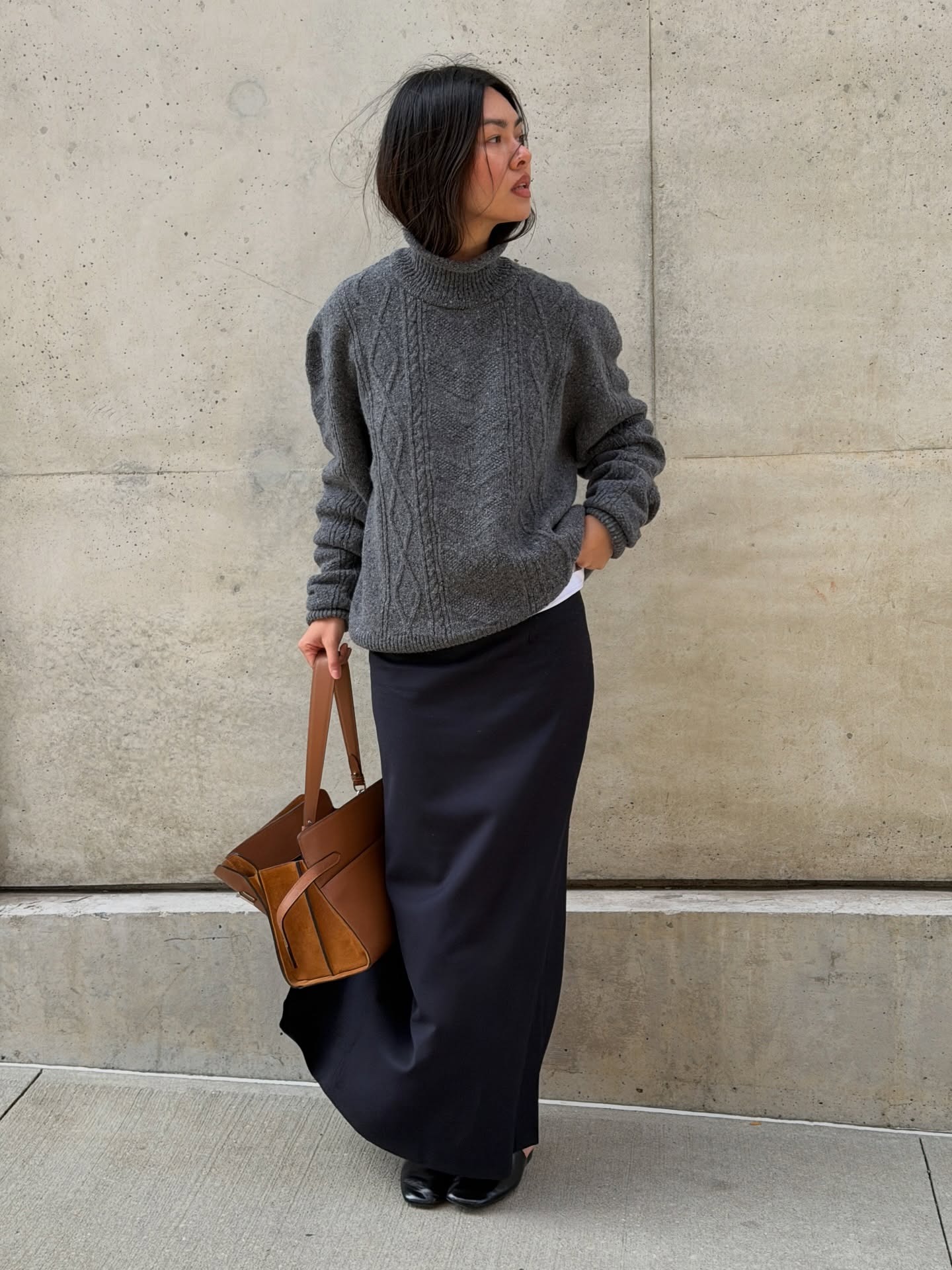 Mimi wears a gray sweater, black pencil skirt, and tan handbag.