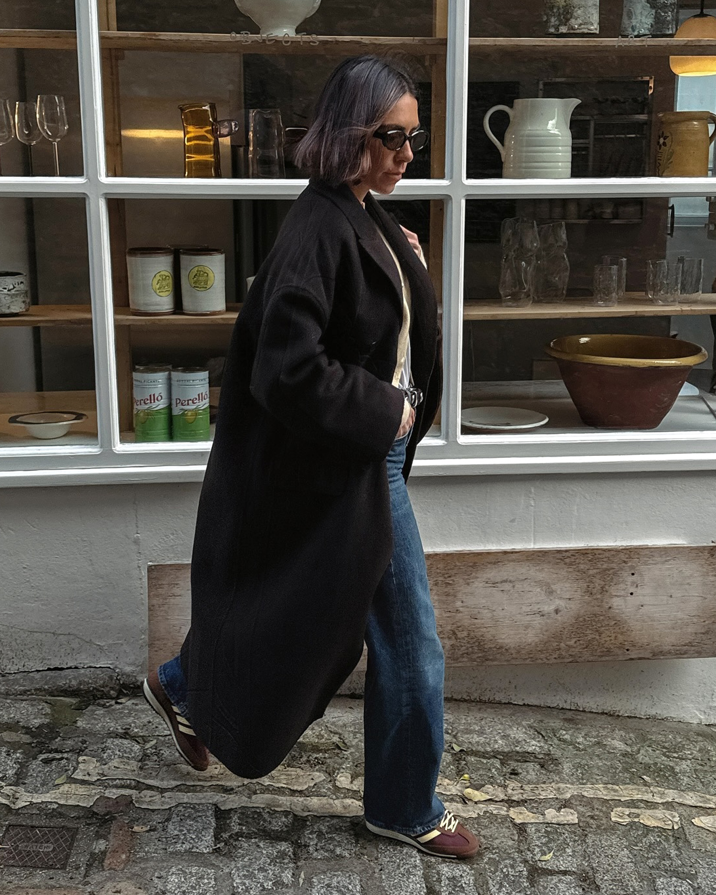 British style influencer Jessica Skye walking a cobblestone street in Cornwall wearing a long brown coat, a studded black belt, relaxed jeans, and brown Adidas SL 72 sneakers