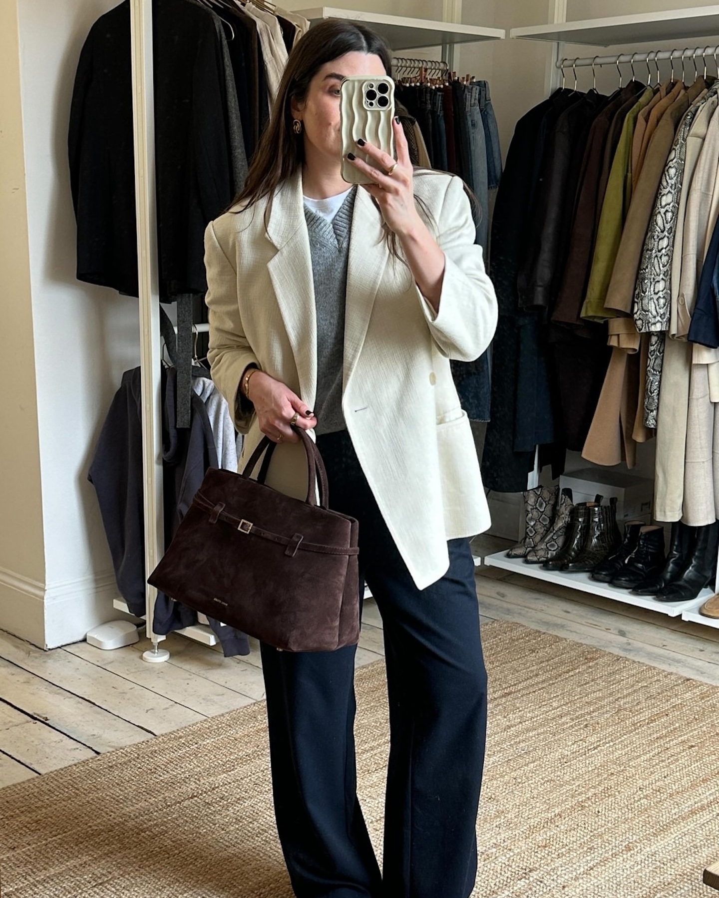 British style influencer Anna Howard poses for a mirror selfie wearing an ivory blazer, gray v-neck sweater, brown suede tote bag, and wide-leg jeans