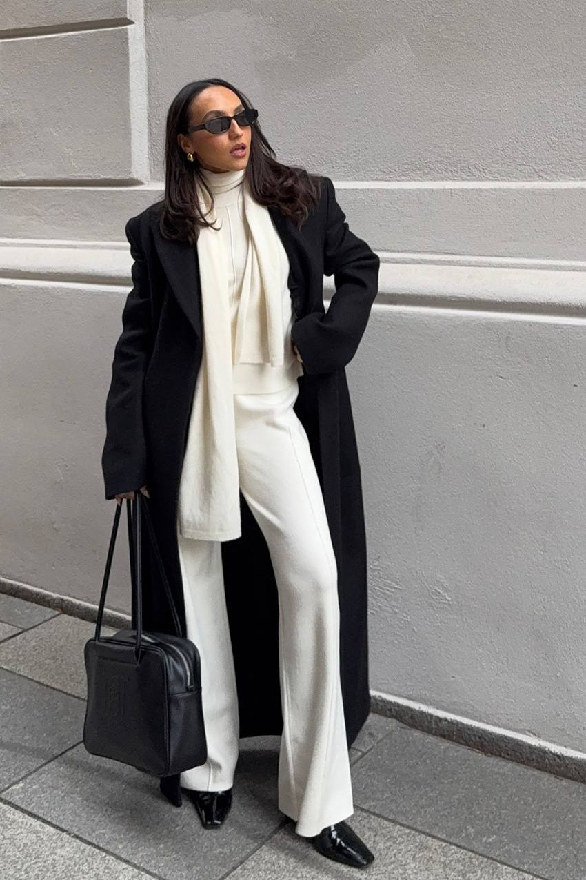 A woman standing on the sidewalk wearing a long black coat over a white sweater with an attached scarf, ivory cashmere pants, black pointed ankle boots, black sunglasses, gold hoop earrings, and a black bowler bag.