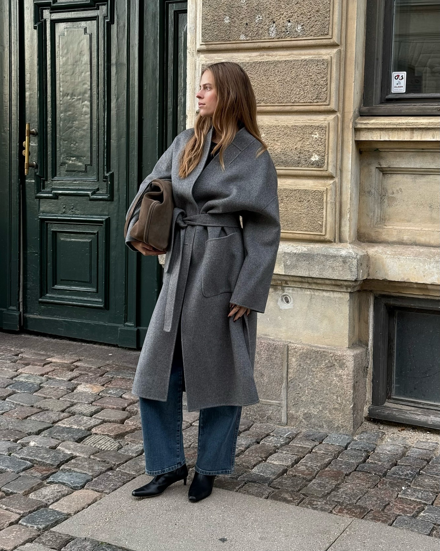 Danish fashion creative Clara Dyrhauge wearing a chic fall and winter outfit with a gray belted coat, a suede bag, jeans, and the black kitten-heel boots trend for 2025